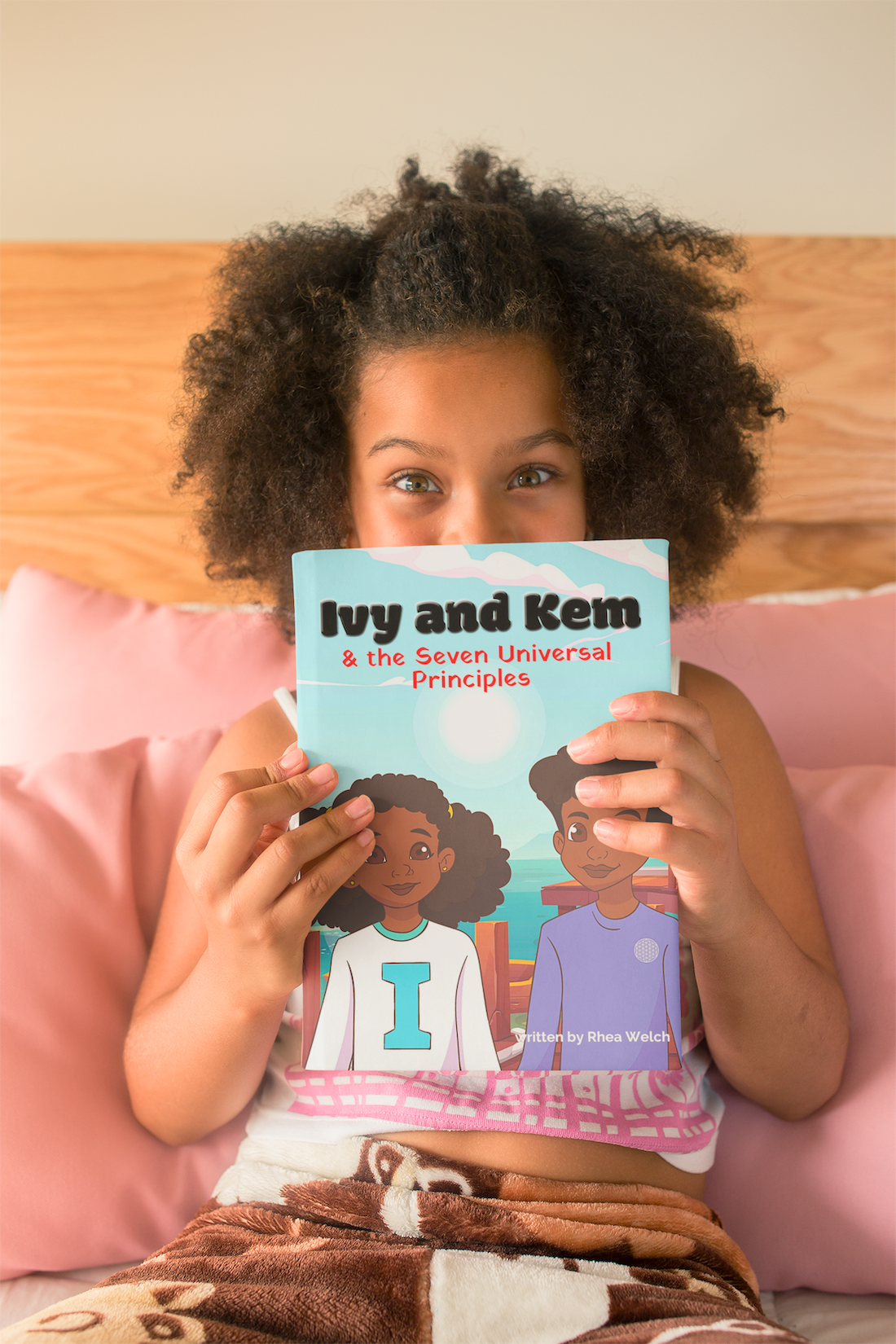 A young girl holds a book titled 'Ivy and Kem & the Seven Universal Principles' in front of her face while sitting on a bed with pink pillows.