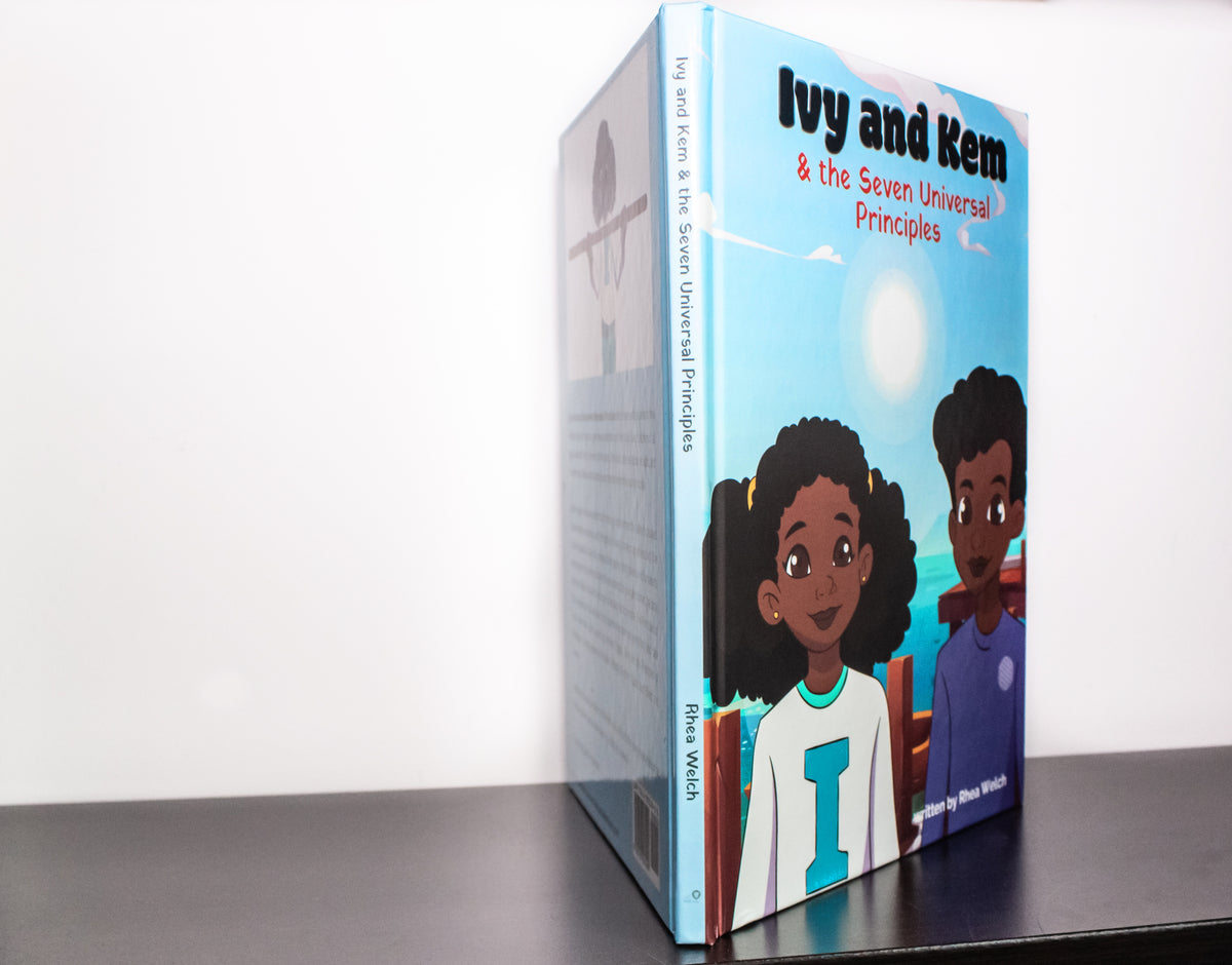A book titled 'Ivy and Kem & the Seven Universal Principles' standing upright on a dark surface against a plain white wall. The cover features illustrated children and a bright blue sky background.