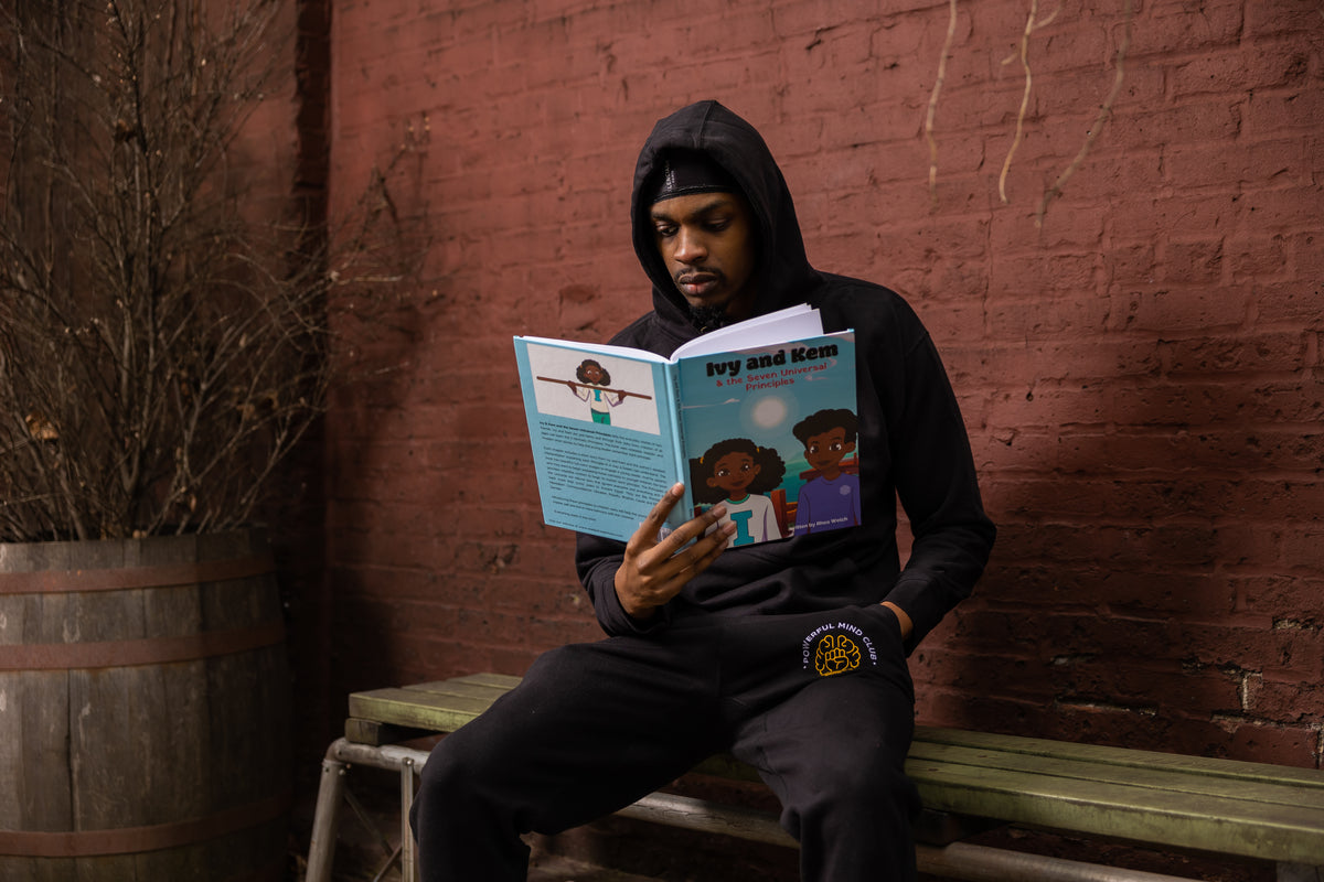 A man in a Powerful Mind Club black hoodie and sweatpants sits on a wooden bench against a red brick wall, reading the book titled "Ivy and Kem & the Seven Universal Principles".
